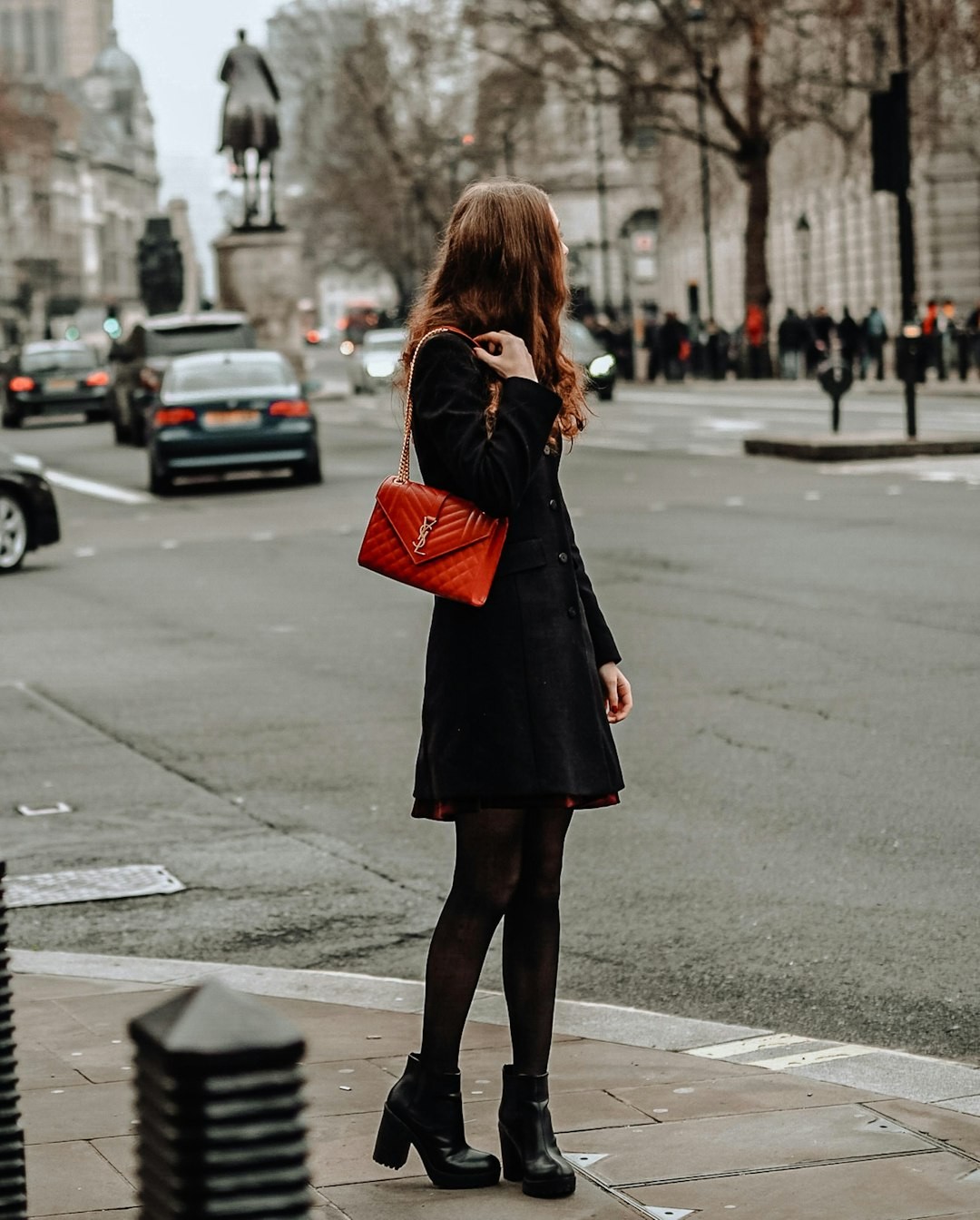 woman in black coat and red leather sling bag standing on sidewalk during daytime
